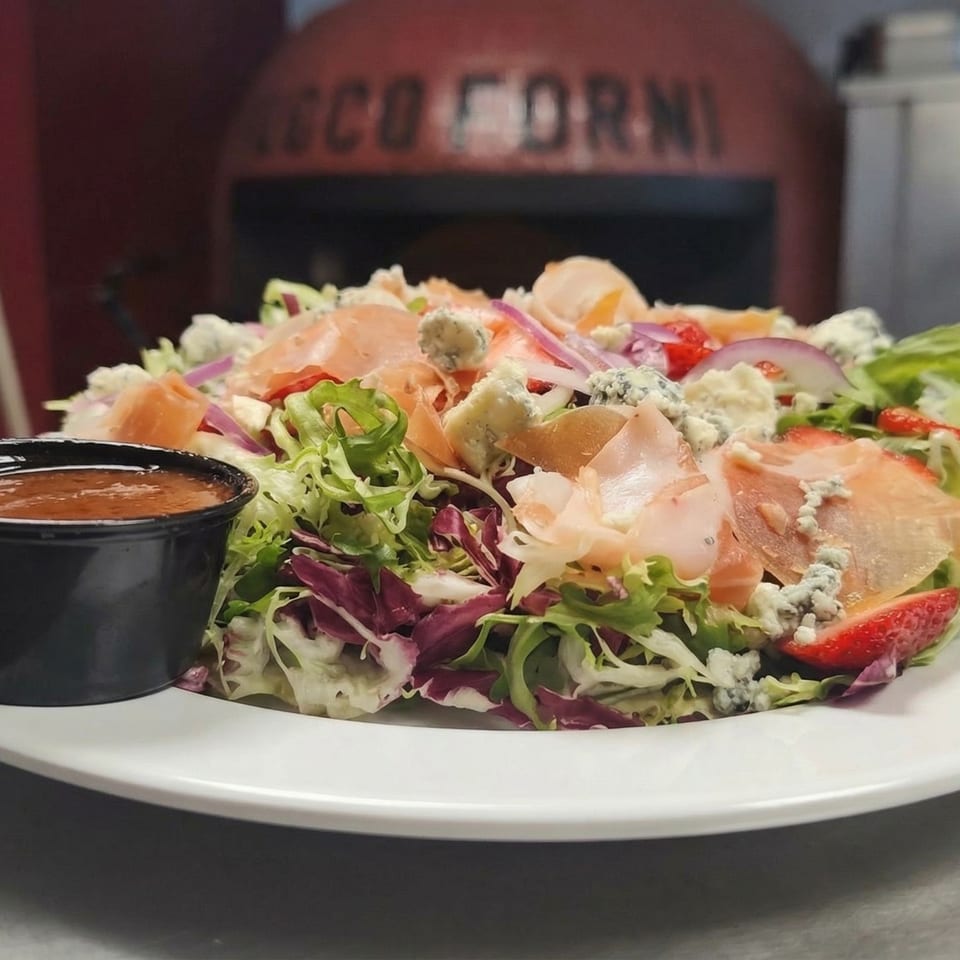 Fresh salad with prosciutto, blue cheese, strawberries, and red onion, with a pizza oven in the background.