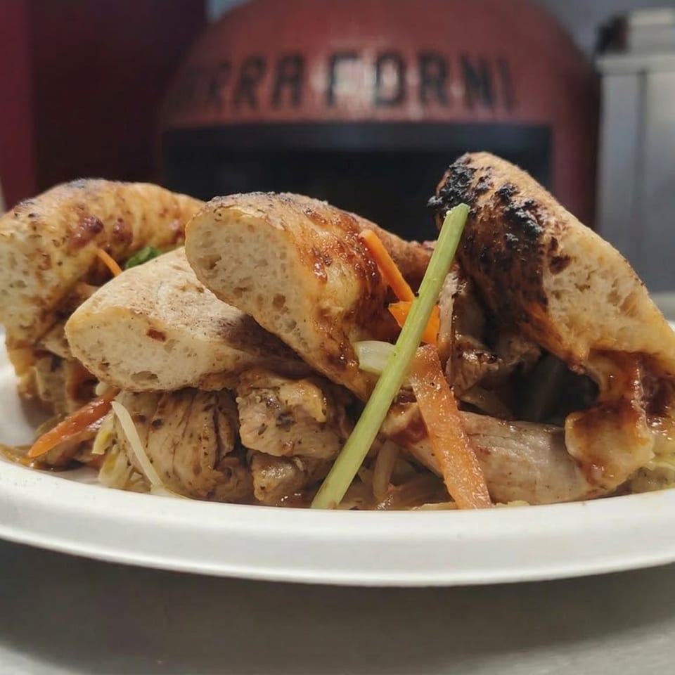 Grilled chicken and vegetables served on toasted baguette slices, with a pizza oven in the background.