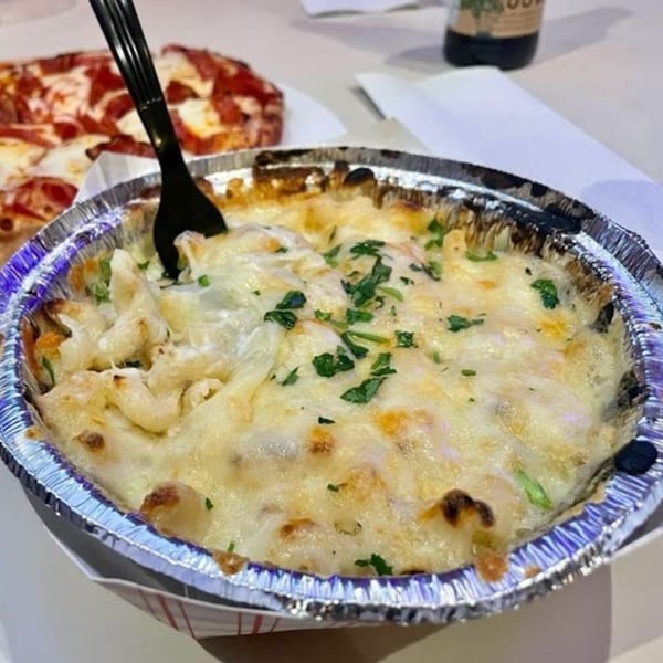 Baked mac and cheese in a foil container with a fork, and a pizza in the background.