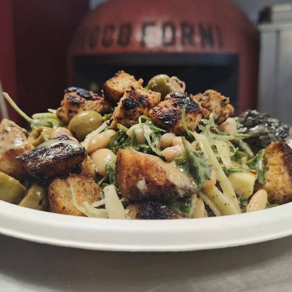 A close-up of a salad with grilled chicken, olives, white beans, and greens, in front of a pizza oven.