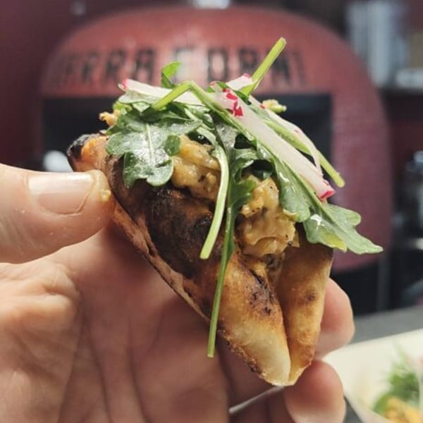Hand holding a folded pizza crust taco with arugula and radishes, a pizza oven in the background.