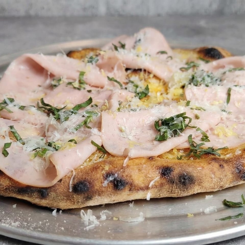 Fire on the Lake pizza with mortadella, grated cheese, and fresh herbs on a metal tray.