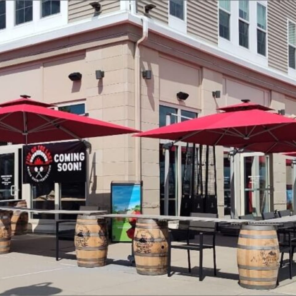 Fire on the Lake pizzeria exterior with red umbrellas, barrel tables, and a "Coming Soon!" banner.